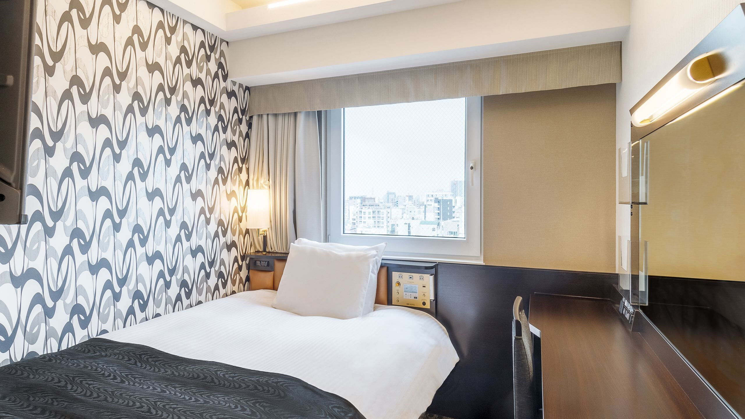 A hotel room with a bed, a nightstand, a lamp, a window with a city view, and a wall with a patterned design.