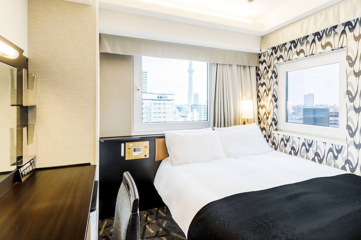 A hotel room with a bed, a desk, a chair, a window with a city view, and a control panel on the wall.