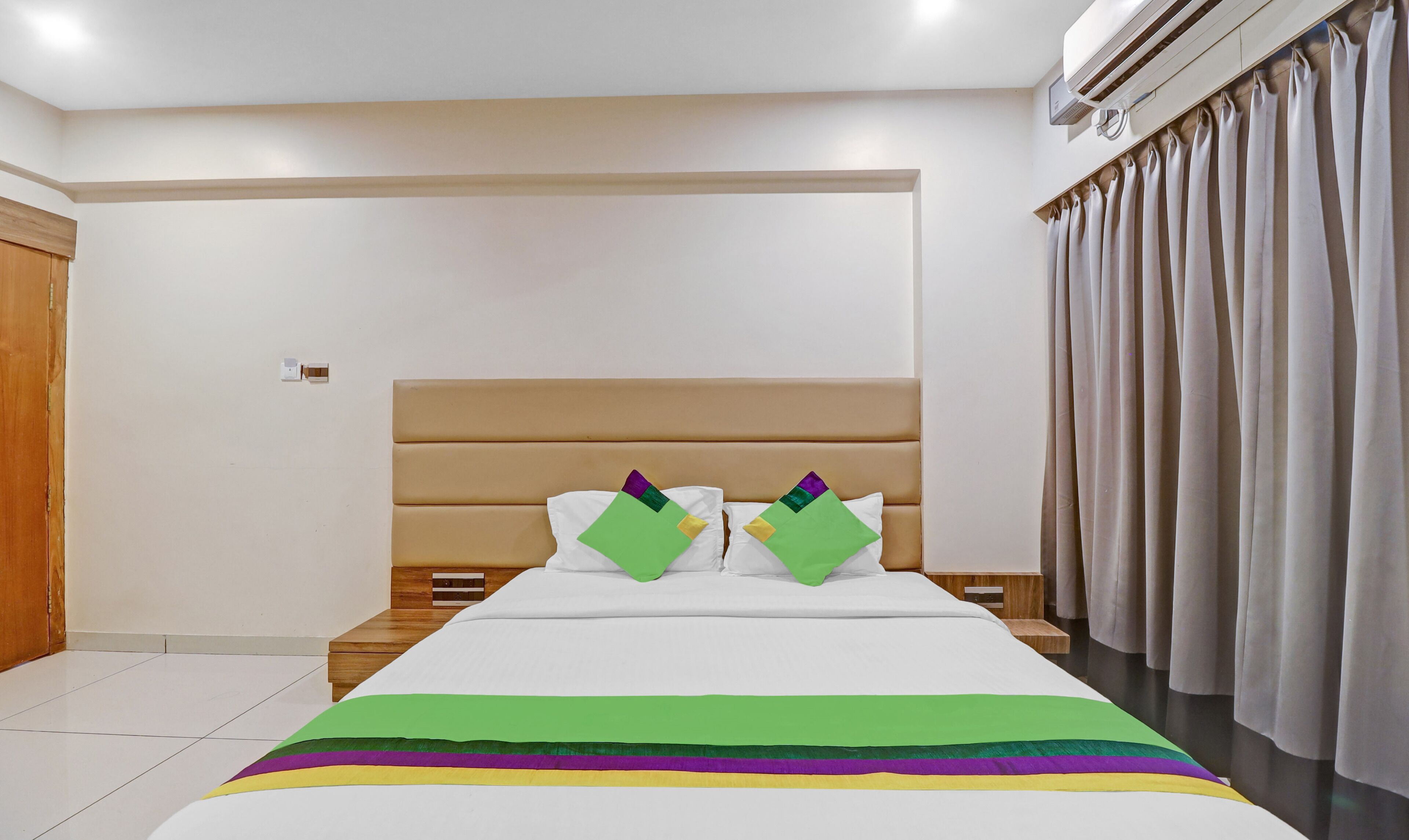 Quarto de hotel com uma cama grande, cabeceira de madeira e uma janela com cortinas.