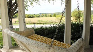 A wicker hammock swing with a yellow cushion, hanging from a porch with a view of a green field and trees. - Entire farmhouse in the country (Winterset)