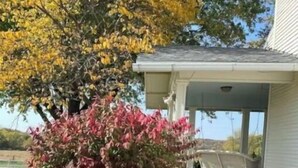 A covered porch with a concrete floor, a white building in the background, and a variety of plants including a bush with red leaves. - Entire farmhouse in the country (Winterset)