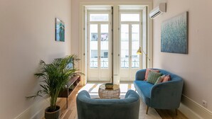 Una sala de estar con un sofá azul, una mesita redonda, una planta en maceta y un amplio ventanal con vistas a balcones.
