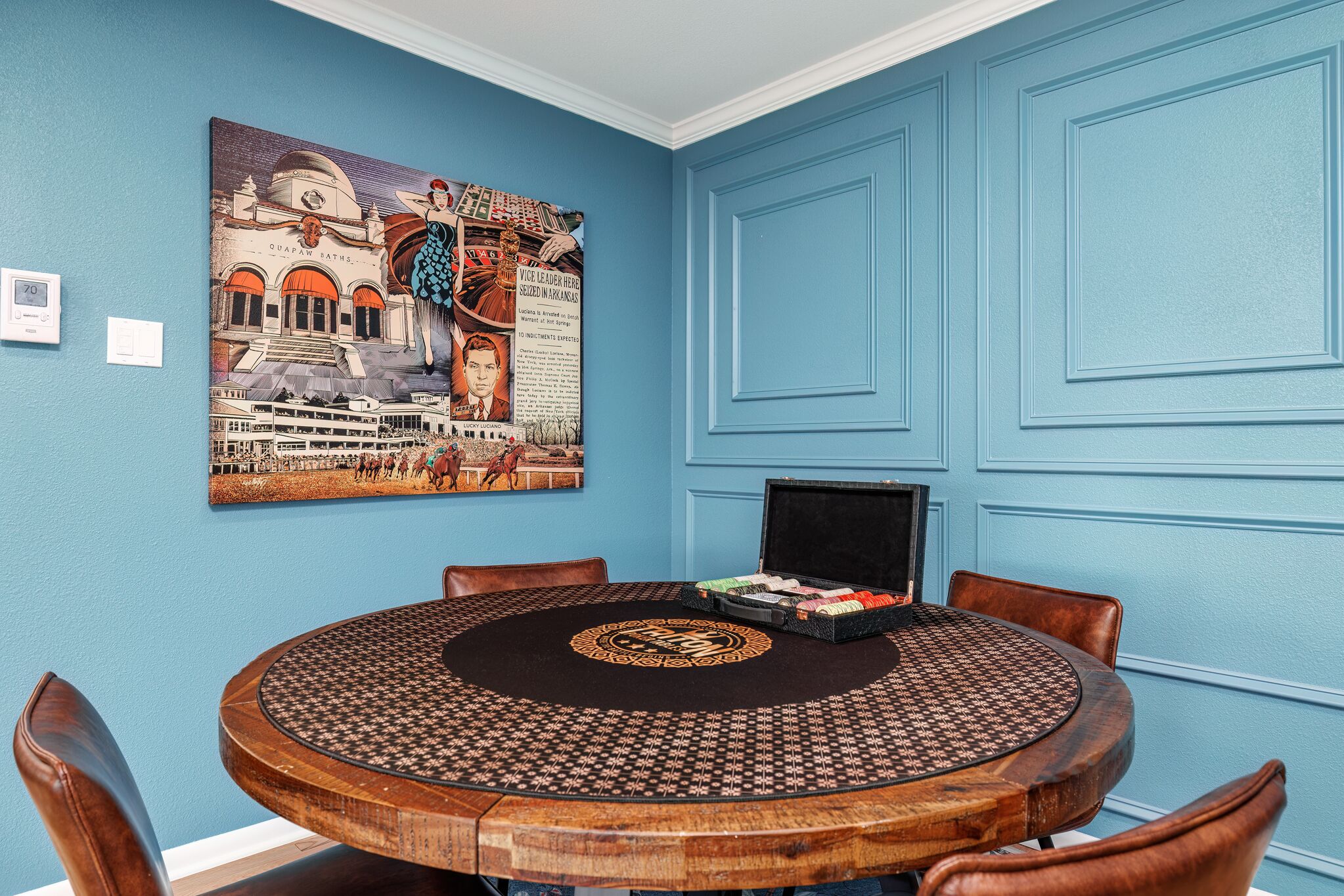 A round wooden table with a patterned tablecloth, surrounded by leather chairs, in a room with blue walls and a large historical painting.