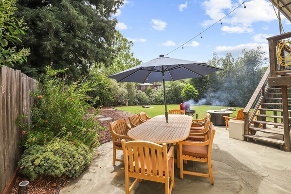 A patio with a wooden table and chairs, an umbrella, and a barbecue grill.
