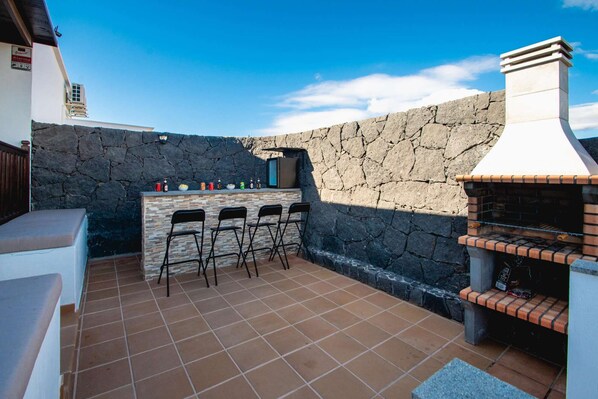 An outdoor patio with a stone wall, a barbecue grill, and a bar area with chairs and bottles.