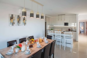 A modern dining area with a wooden table set for breakfast, white chairs, and pendant lighting.