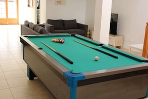 A pool table with balls and cues, a sofa, and a television in a room with tiled flooring.