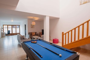 A pool table in a living room with a sofa and a chair, a staircase, and a door leading to another room.