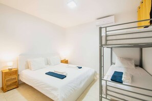A hotel room with a single bed, a bunk bed, a bedside table with a lamp, and an air conditioning unit on the wall.