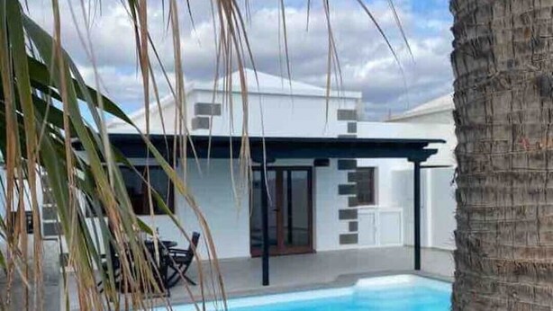 Un área de piscina moderna al aire libre con zona de estar integrada, una palmera y un cielo azul despejado.