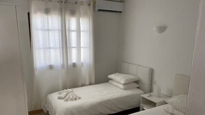 Una habitación con dos camas individuales, una ventana con cortinas translúcidas, un equipo de aire acondicionado y una mesita de noche con lámpara.