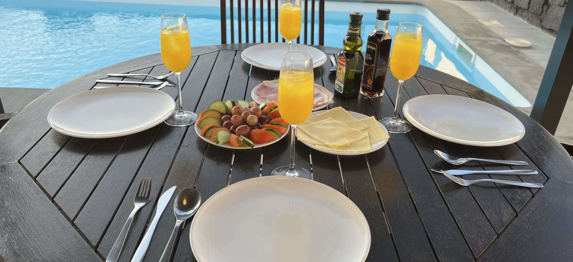 A table set for two with plates, cutlery, a glass of orange juice, a bottle of olive oil, and a bottle of balsamic vinegar.