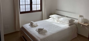 A clean, well-lit hotel room with a neatly made bed, a bedside table with a lamp, and a window with curtains.