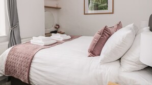 A neatly made bed with white and pink bedding, a framed picture of a forest on the wall, and a small bedside table with a mirror and a vase of flowers. - Ermysted's Cottage (Skipton)
