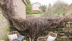 A patio with wicker chairs and a table, a stone wall, and a garden in the background. - Ermysted's Cottage (Skipton)