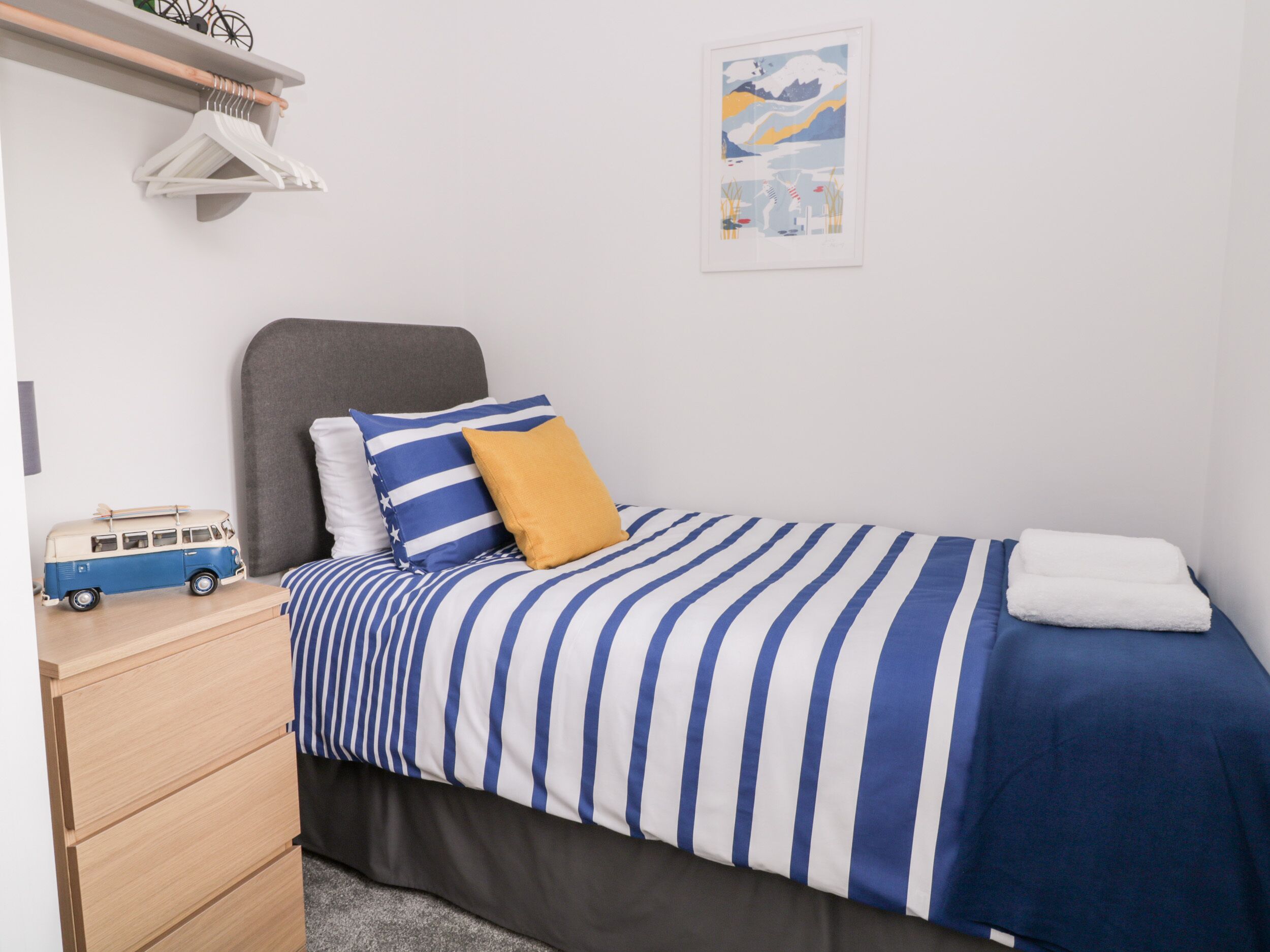 Un dormitorio ordenado con cama, una mesita de noche pequeña, un estante con ropa y un cuadro colgado en la pared.