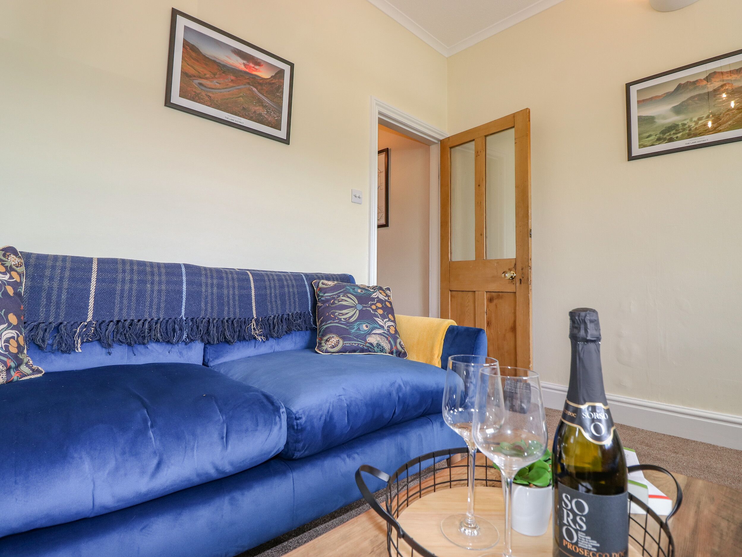 A blue sofa with a patterned throw, a wine bottle, and glasses on a tray, with framed pictures on the walls.