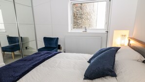 A neatly made bed with a white and blue duvet, a blue chair, and a view of a stone wall through a window. - Harbour Hideaway (Looe)