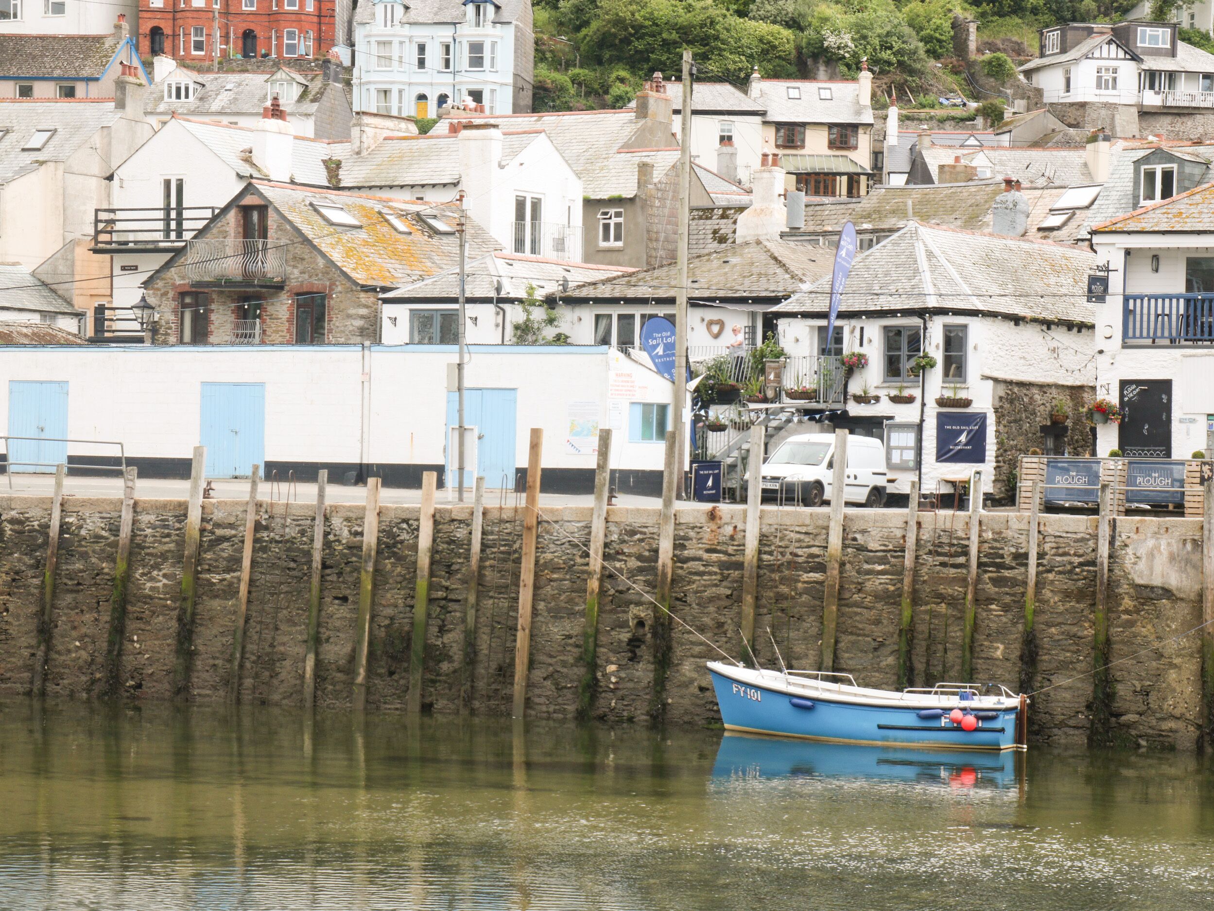 A coastal town with a row of white buildings, a blue boat docked at the pier, and a stone wall in the foreground.