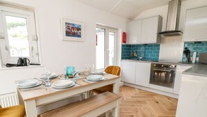 A modern kitchen with a wooden dining table set for four, white cabinets, a stainless steel range hood, and a view of the street through the window. - Harbour Hideaway (Looe)