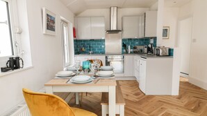 A modern kitchen with white cabinets, a teal backsplash, and a dining area with a wooden table and a mustard-colored chair. - Harbour Hideaway (Looe)