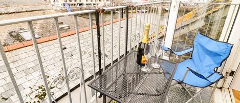 Un balcon avec une table en métal, deux chaises bleues, une bouteille de champagne et un verre.