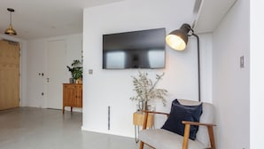 Een moderne woonkamer met een flatscreen televisie aan de muur, een houten bijzettafeltje met een bloemenvaas, en een lichtkleurige bank met een donkere kussen.