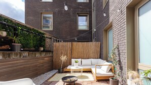 Een modern terras buiten met een houten terras, een witte bank, een ronde tafel en een vuurkuil.