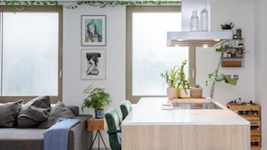Een moderne woonkamer met een grijze bank, een blauwe plaids en een groene fauteuil. Er is een keukenblok met krukken en een potplant.