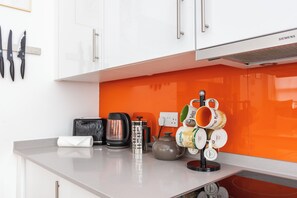 Een keuken met witte kastjes, een oranje spatwand, een koffiezetapparaat, een waterkoker en een muggenhouder met meerdere mokken.