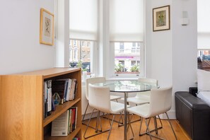Een moderne leefruimte met een glazen tafel, omringd door witte stoelen, een houten boekenkast en een zwarte leren bank.