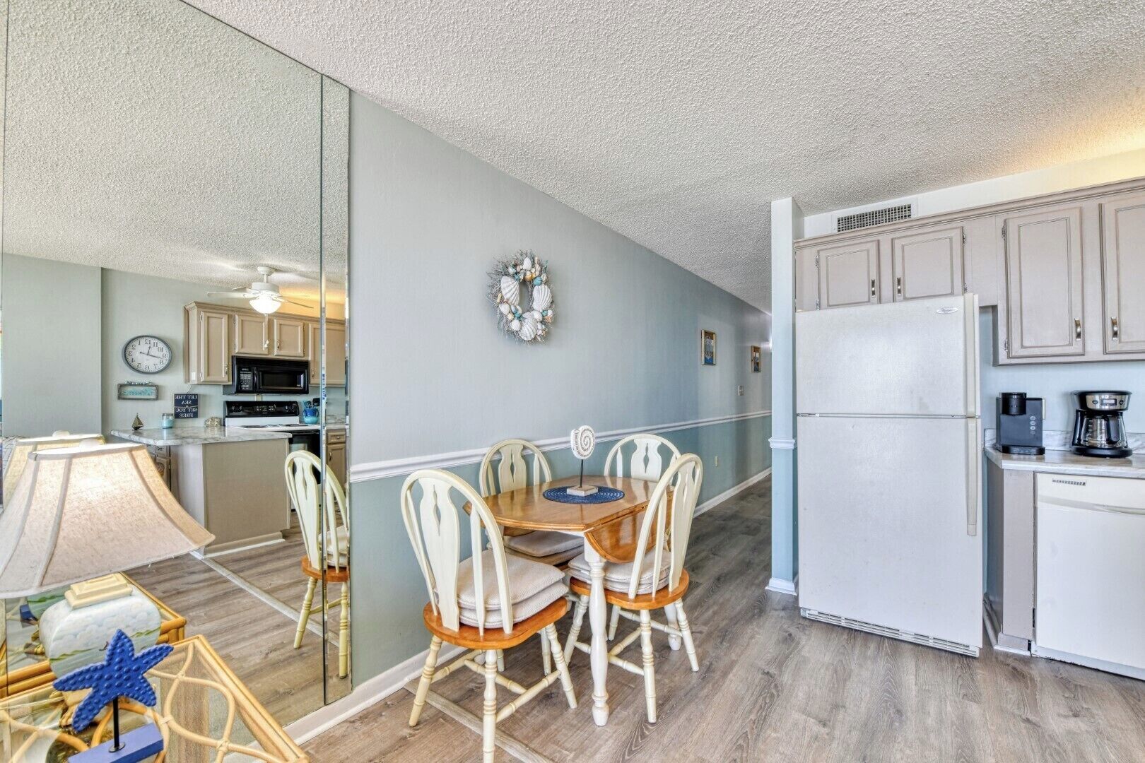 A compact kitchen with white cabinets, a refrigerator, and a microwave. There is a dining area with a table and chairs, and a decorative wreath on the wall.
