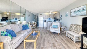 Ein geräumiges Wohnzimmer mit einer Couch, Sesseln und einem Couchtisch. Im Hintergrund ist eine Küche mit Essbereich zu sehen.