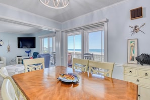 A dining area with a wooden table, chairs, a television, and a view of the beach.