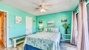 Une chambre avec un mur de couleur turquoise, un lit recouvert d’une courtepointe à motifs bleus et blancs, un ventilateur de plafond et des œuvres d’art accrochées aux murs.