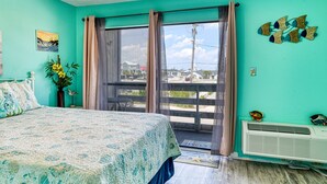 Une pièce aux murs de couleur turquoise, un lit recouvert d’une couvre-lit à motifs floraux, une fenêtre avec des rideaux, une petite table sur laquelle est posé un vase de fleurs et un climatiseur fixé au mur.