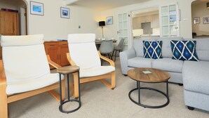 A living room with a grey sofa, two white armchairs, a wooden coffee table, and a small round table. There is a kitchen visible through an open door in the background.