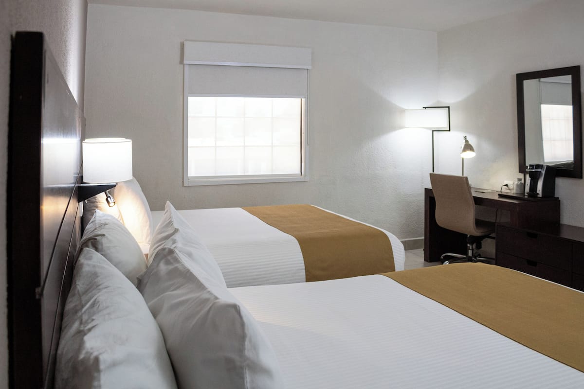 Quarto de hotel com duas camas, uma escrivaninha, uma cadeira, um abajur e janela com persianas.
