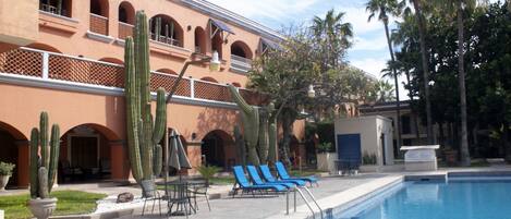 Un área de piscina con sillones de descanso, un jardín de cactus y un edificio con ventanas arqueadas.