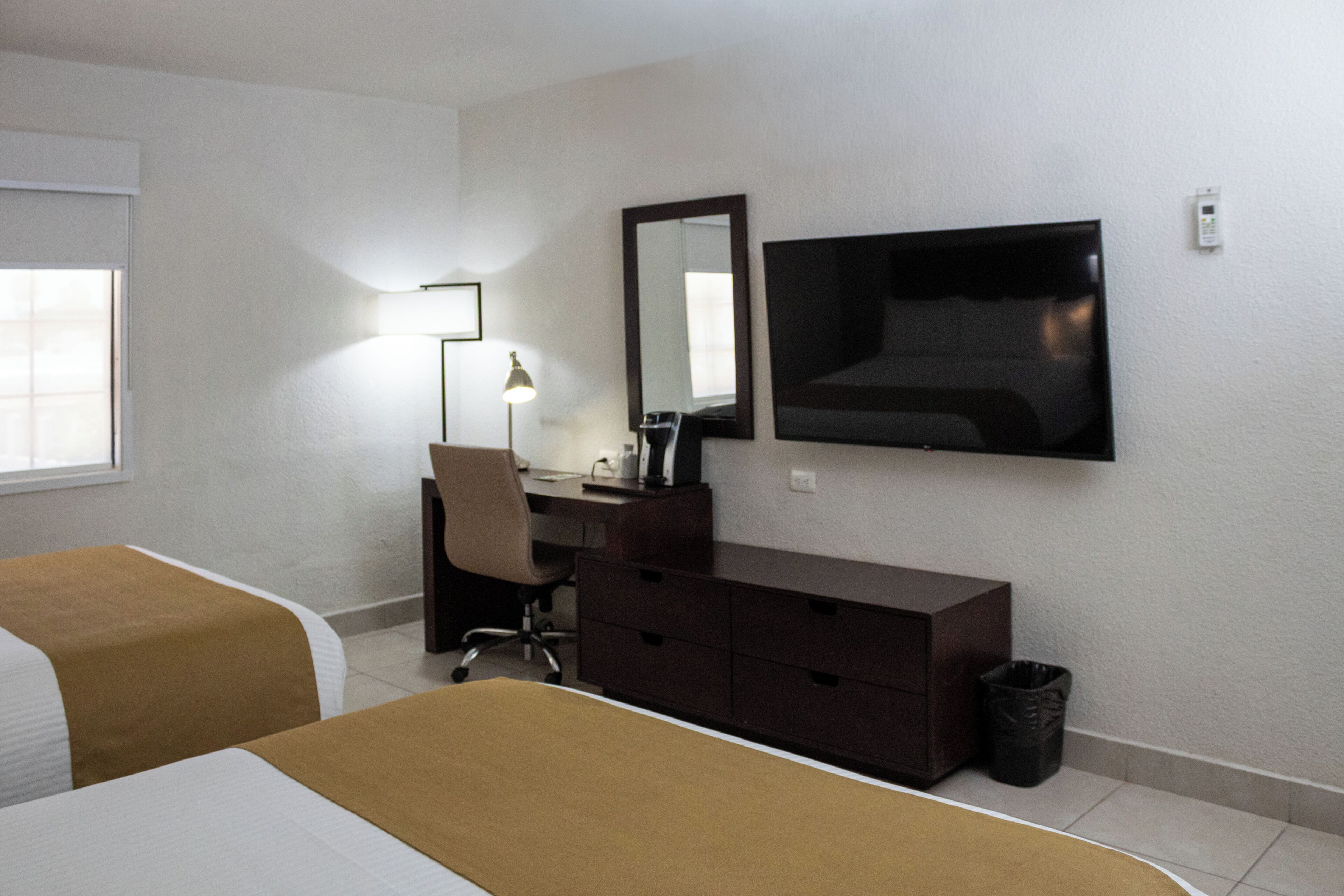 A hotel room with two beds, a desk, a chair, a television, a mirror, and a waste bin.