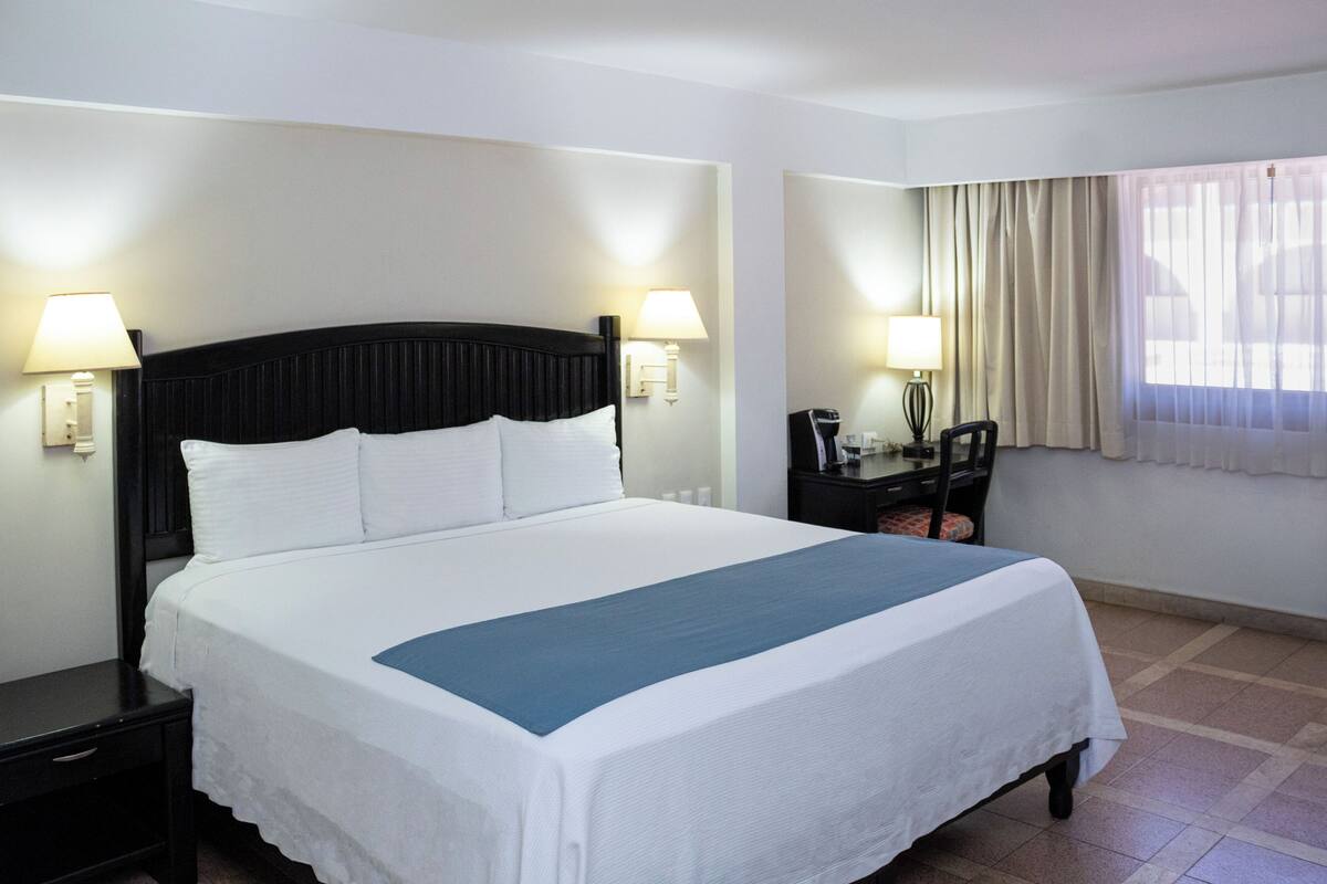 Uma cama bem arrumada com roupa de cama branca e um toque de azul, com duas mesas de cabeceira com abajures, num quarto com design simples e limpo, e uma janela com cortinas.