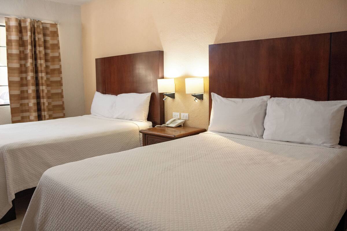 Quarto de hotel com duas camas de solteira, cabeceira de madeira, luminárias laterais e telefone no criado-mudo.