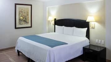 Una cama bien hecha con una colcha blanca y azul, dos lámparas de noche, un cuadro en la pared y una mesita de noche pequeña y negra.