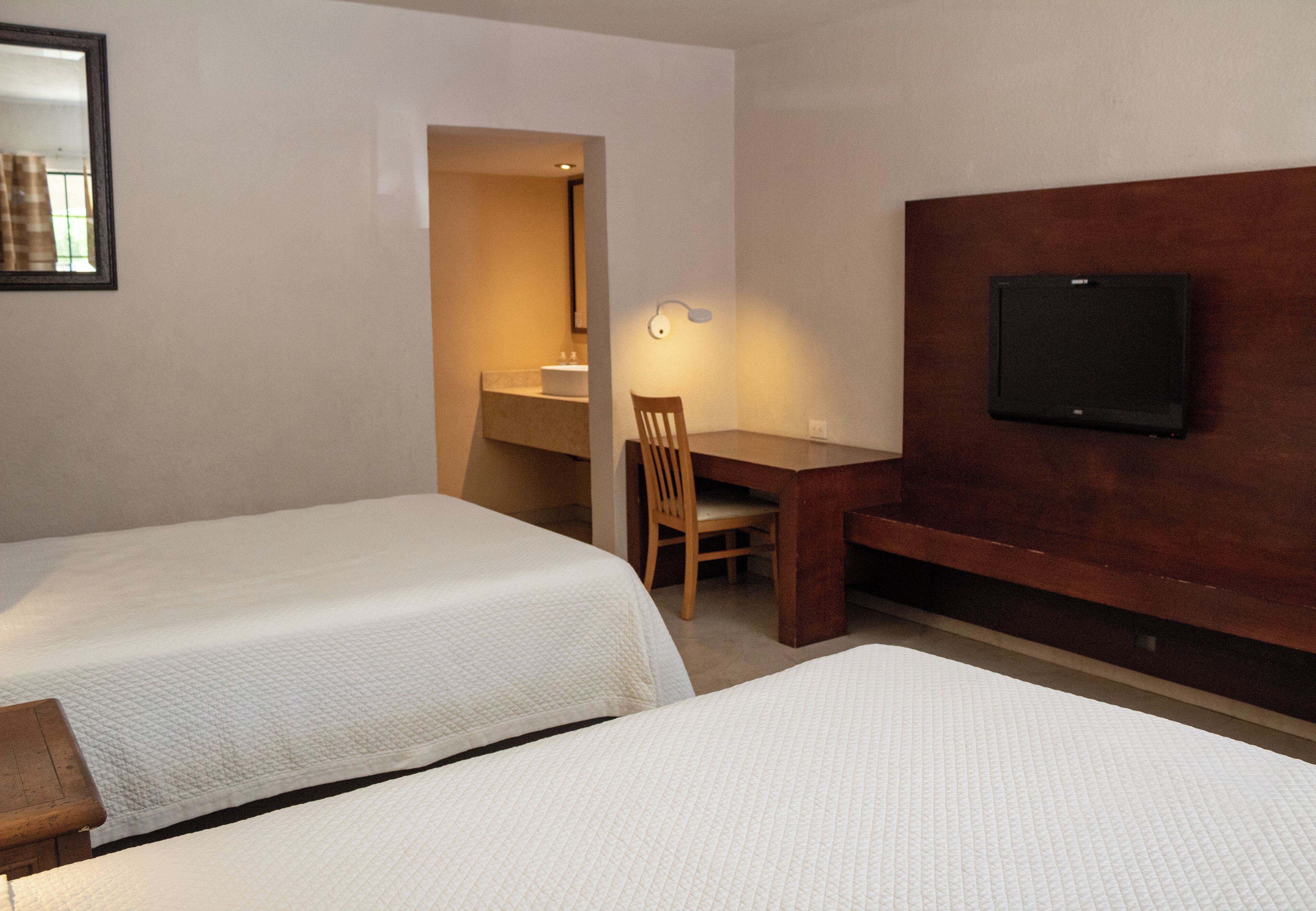 Habitación de hotel con dos camas individuales, un escritorio de madera, una silla, un televisor montado en la pared y un baño visible a través de una puerta abierta.