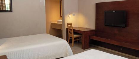 Habitación de hotel con dos camas individuales, un escritorio de madera, una silla, un televisor montado en la pared y un baño visible a través de una puerta abierta.