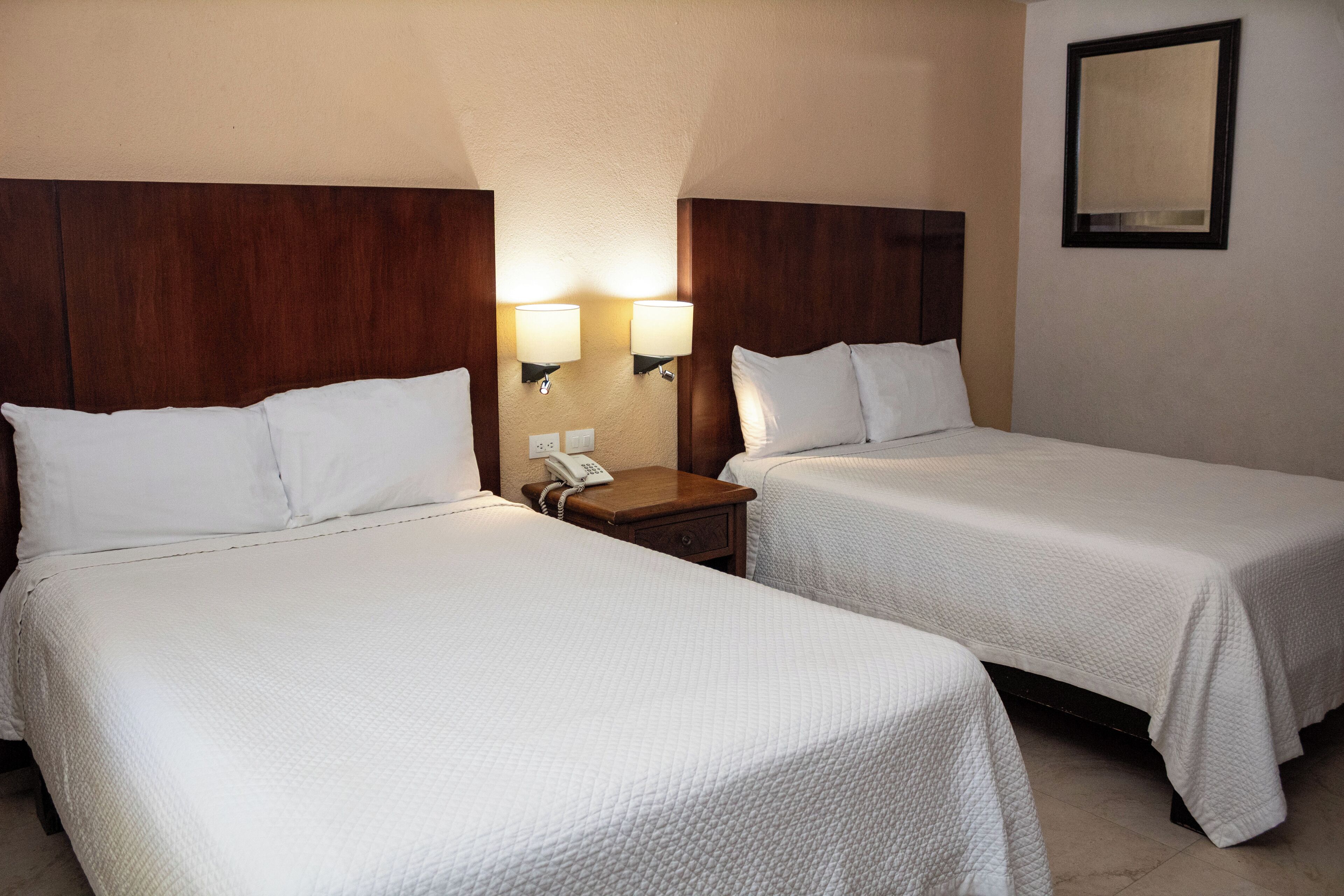 Two single beds with white linens, a wooden headboard, a nightstand with a phone, and wall-mounted lamps.