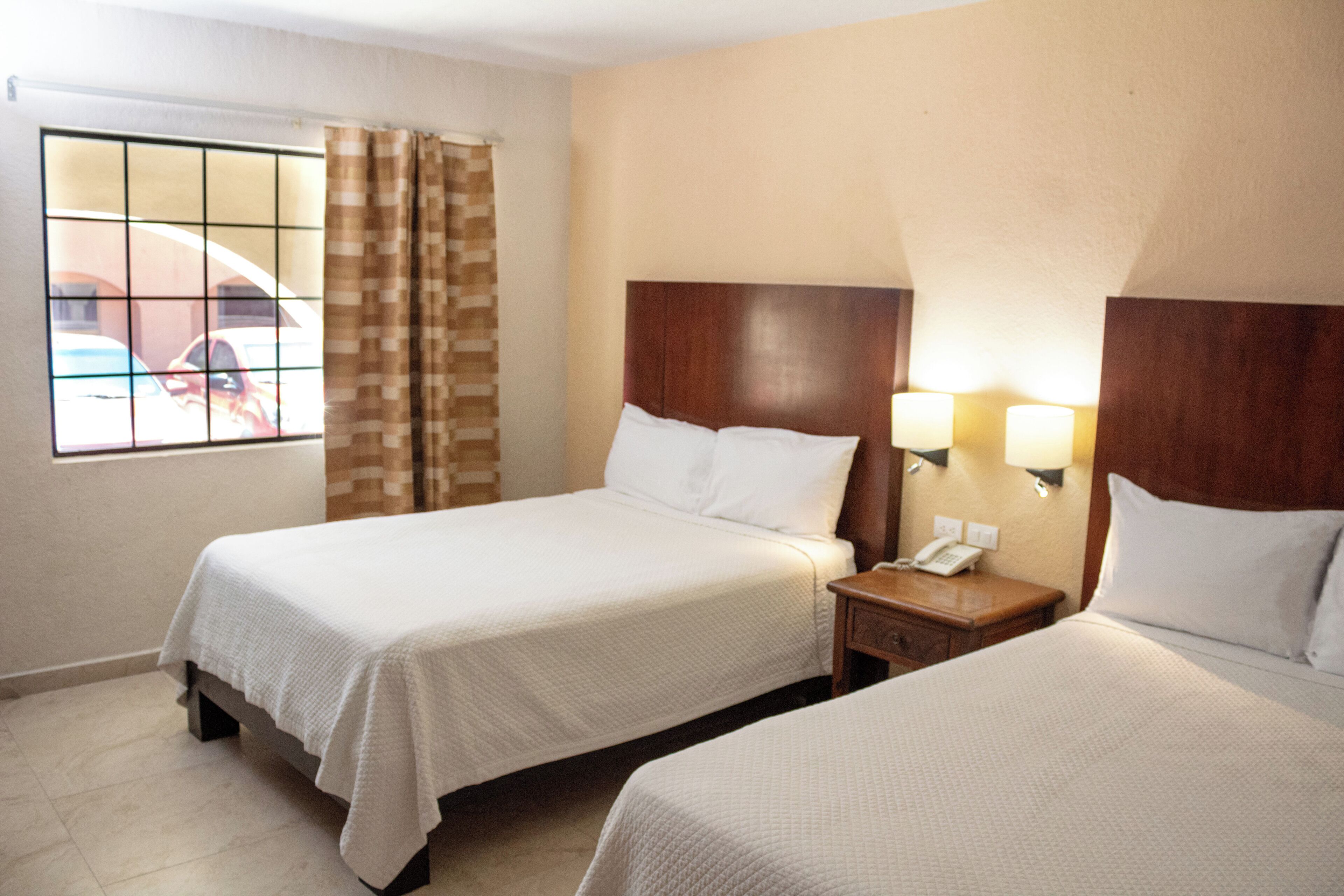 Une chambre d’hôtel avec deux lits, une fenêtre donnant sur les voitures garées et une table de chevet avec un téléphone.