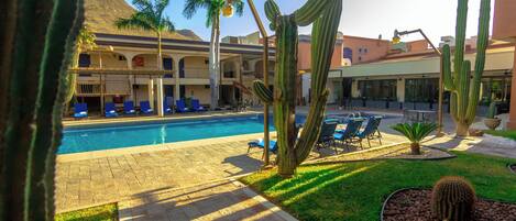 A pool area with lounge chairs, cacti, and a building with arched windows.