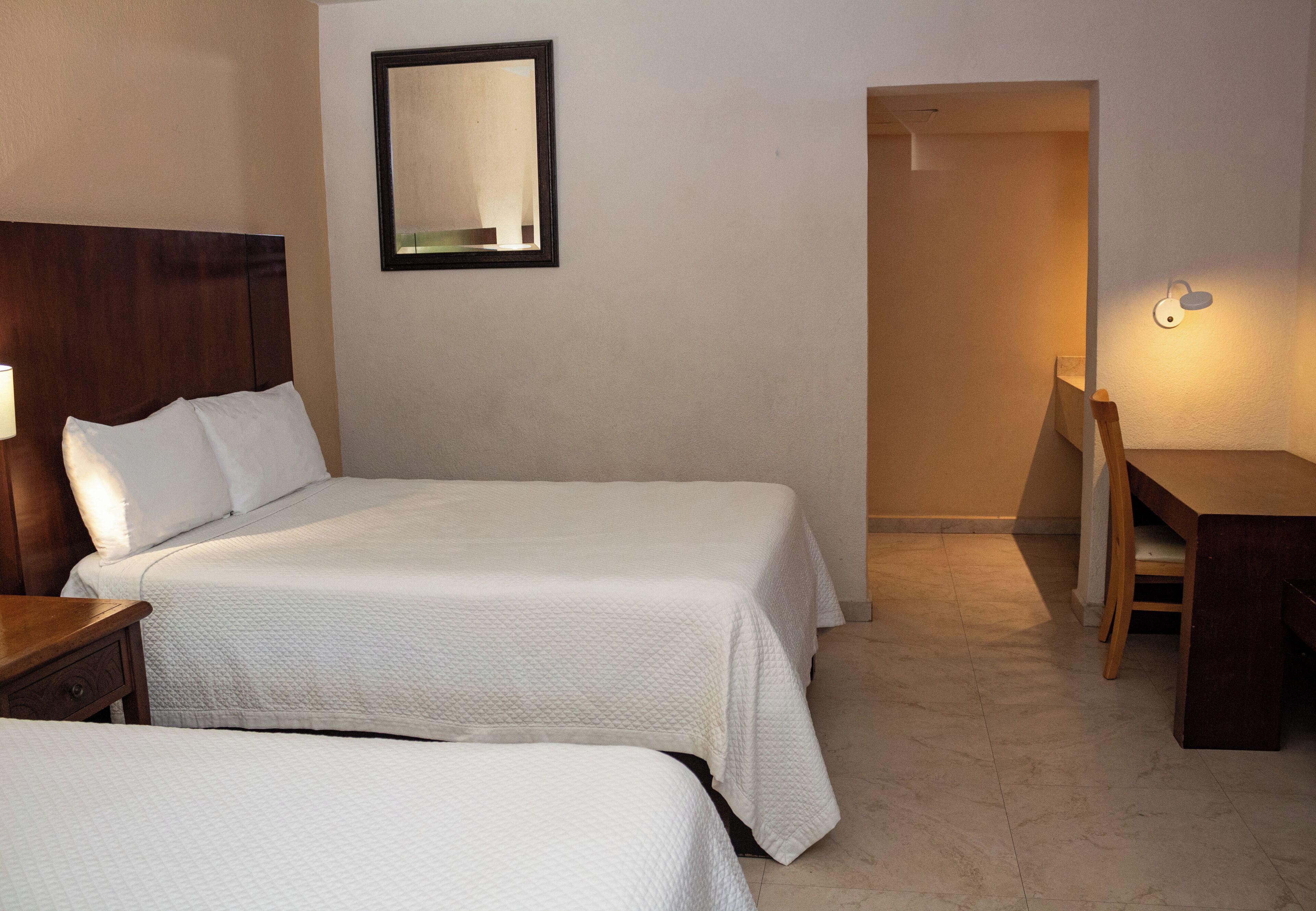 Habitación de hotel con dos camas, un escritorio, una silla, un espejo y una lámpara fijada a la pared.