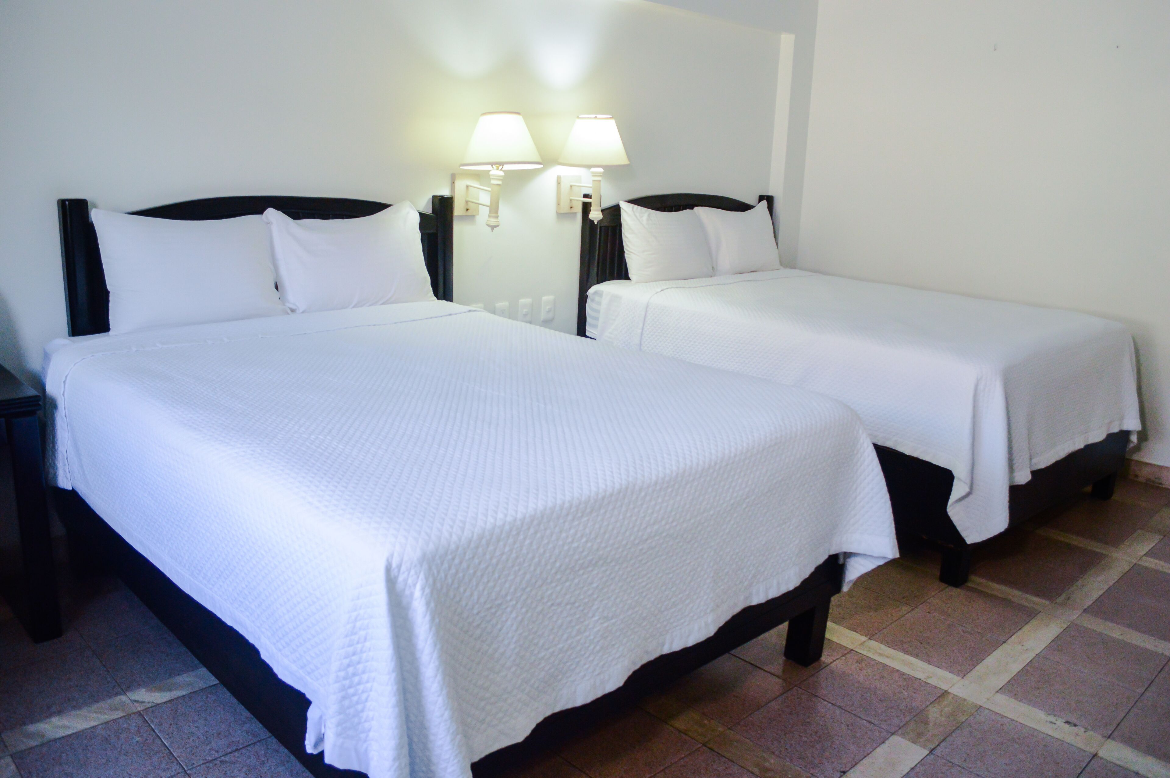 Quarto de hotel com duas camas, cada uma com roupa de cama branca e cabeceiras, e duas luminárias fixadas na parede.